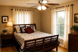 A bedroom with a bed, a nightstand, a lamp, a ceiling fan, a window with curtains, and a plant.