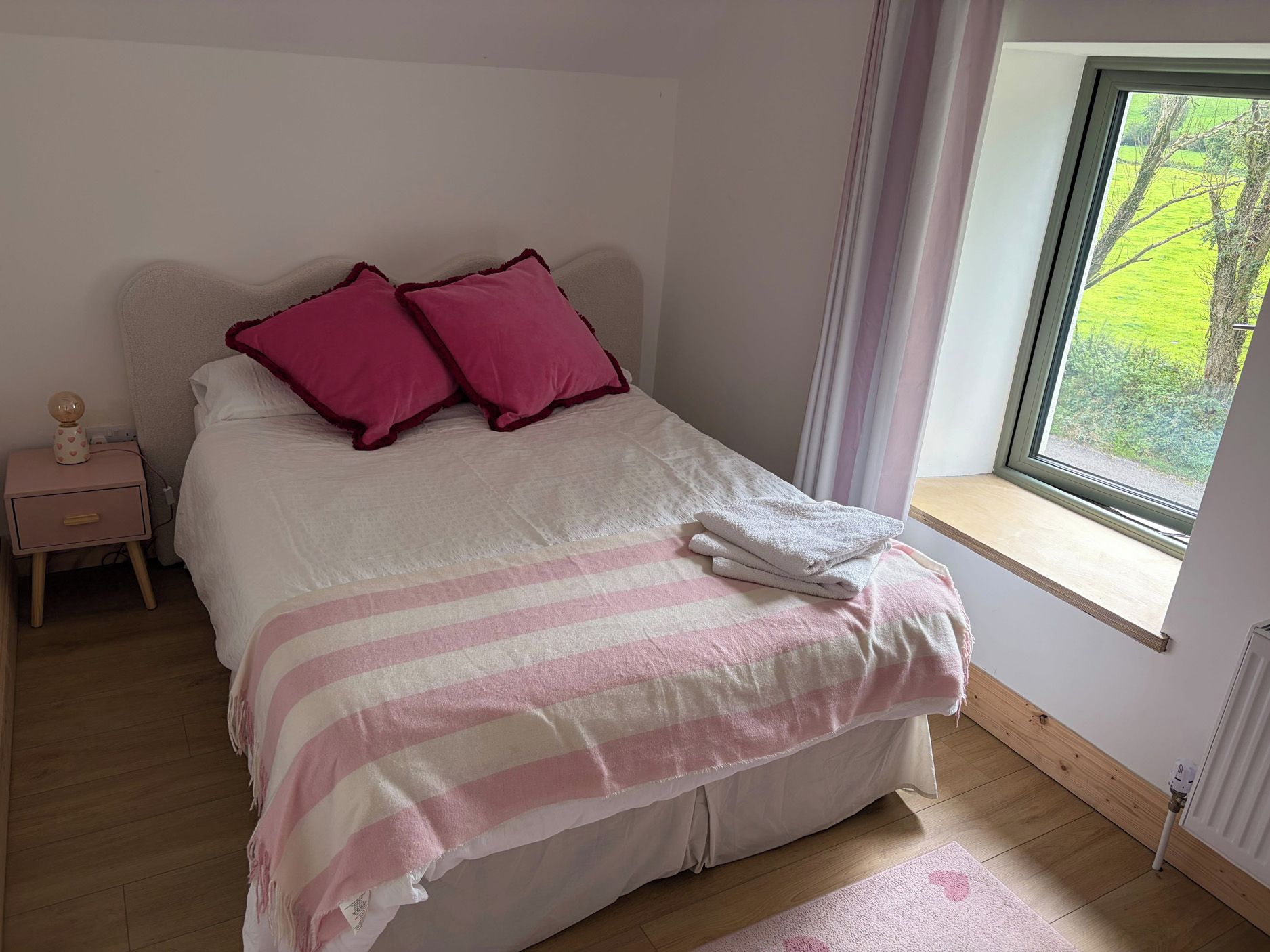 A bedroom with a bed, pillows, a nightstand, a window, and a view of greenery outside.
