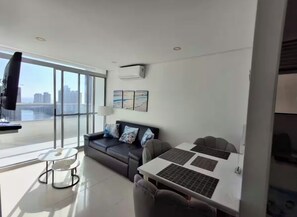 A modern living room with a sofa, a dining table, and a large window with a city view. - Spacious 3-bedroom condo in delightful Cartagena with AC and gym (Cartagena)