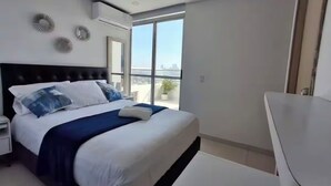 A modern bedroom with a large bed, a nightstand, a white desk, and a view of the city through a large window. - Spacious 3-bedroom condo in delightful Cartagena with AC and gym (Cartagena)