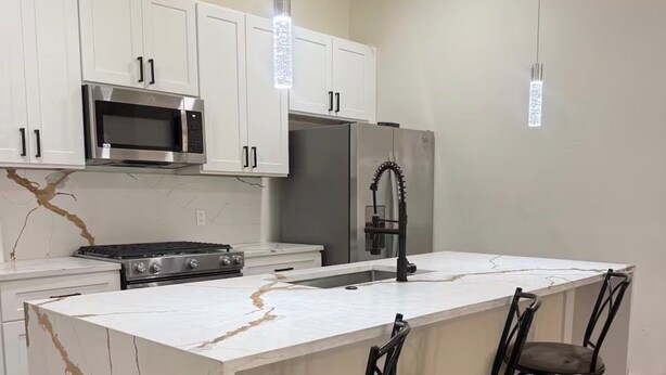 A modern kitchen with white cabinets, a stainless steel microwave, a refrigerator, a gas stove, and a marble island with bar stools. - Fantastic 4BR Houston Chalet Near NRG, TSU, UofH, & Medical Center (Houston)