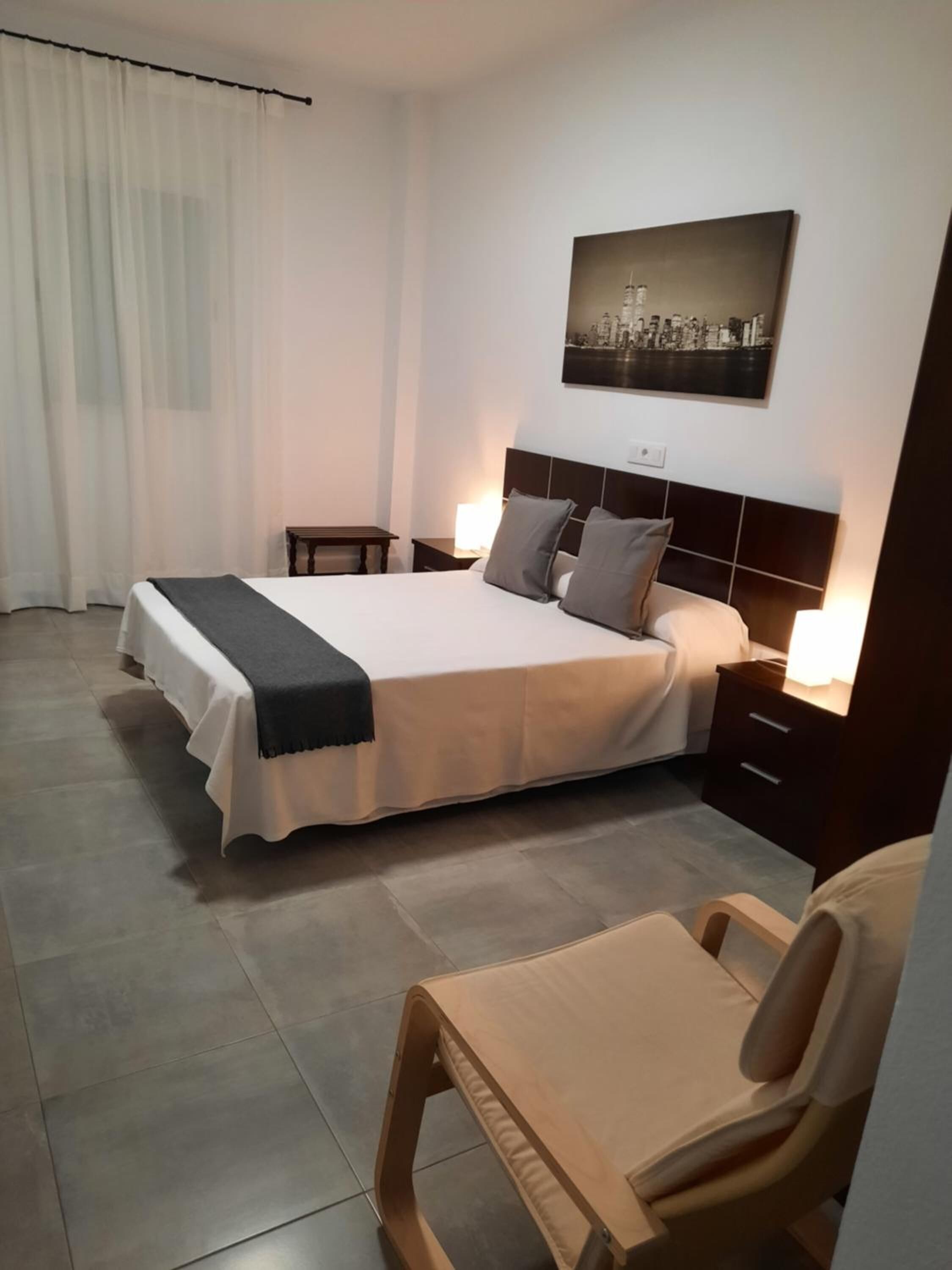 Una habitación de hotel con una cama grande, mesitas de noche, una silla, un cuadro con una vista urbana y una cortina.