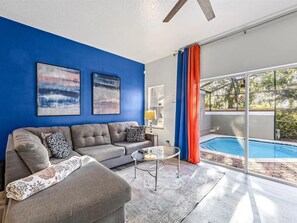 Una sala de estar con pared azul, sofá gris, dos cuadros abstractos, una mesa de centro de vidrio y vista a una piscina.