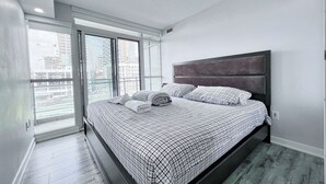 Ein modernes Schlafzimmer mit einem großen Bett, einem grau gepolsterten Kopfteil und Blick auf eine Stadtlandschaft durch raumhohe Fenster.