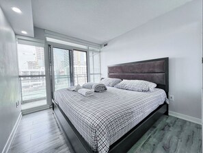Ein modernes Schlafzimmer mit einem großen Bett, einem grau gepolsterten Kopfteil und Blick auf eine Stadtlandschaft durch raumhohe Fenster.