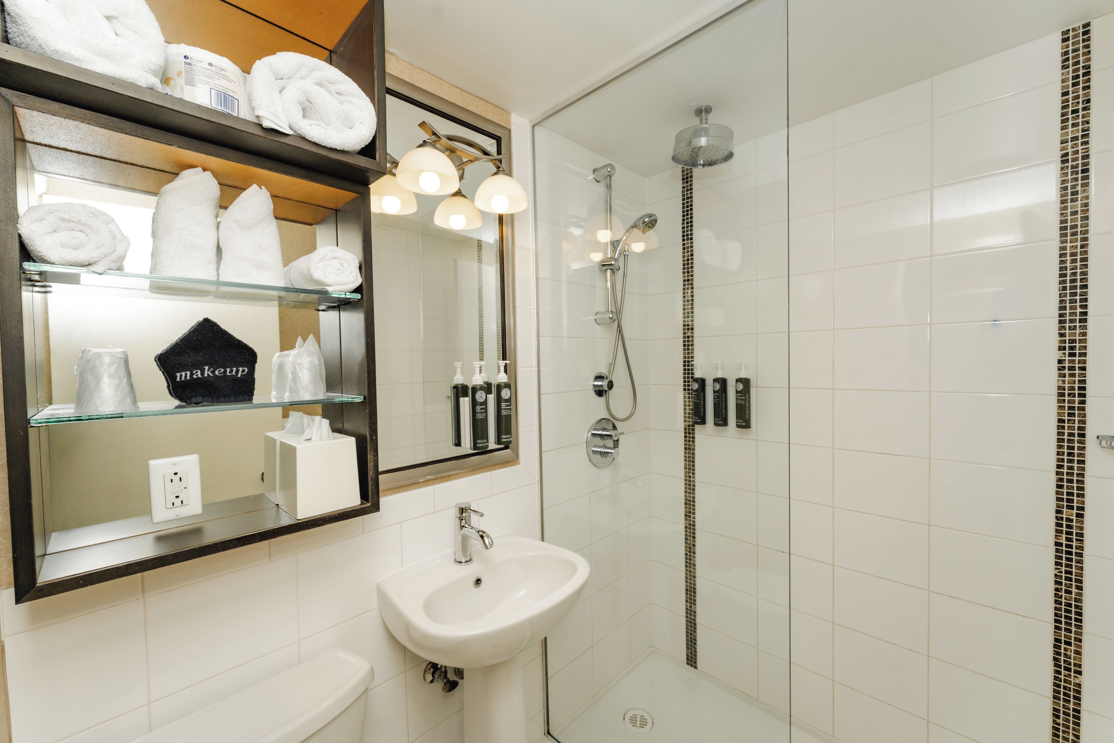 A bathroom with a glass shower enclosure, a sink, a mirror, and shelves with towels and toiletries.