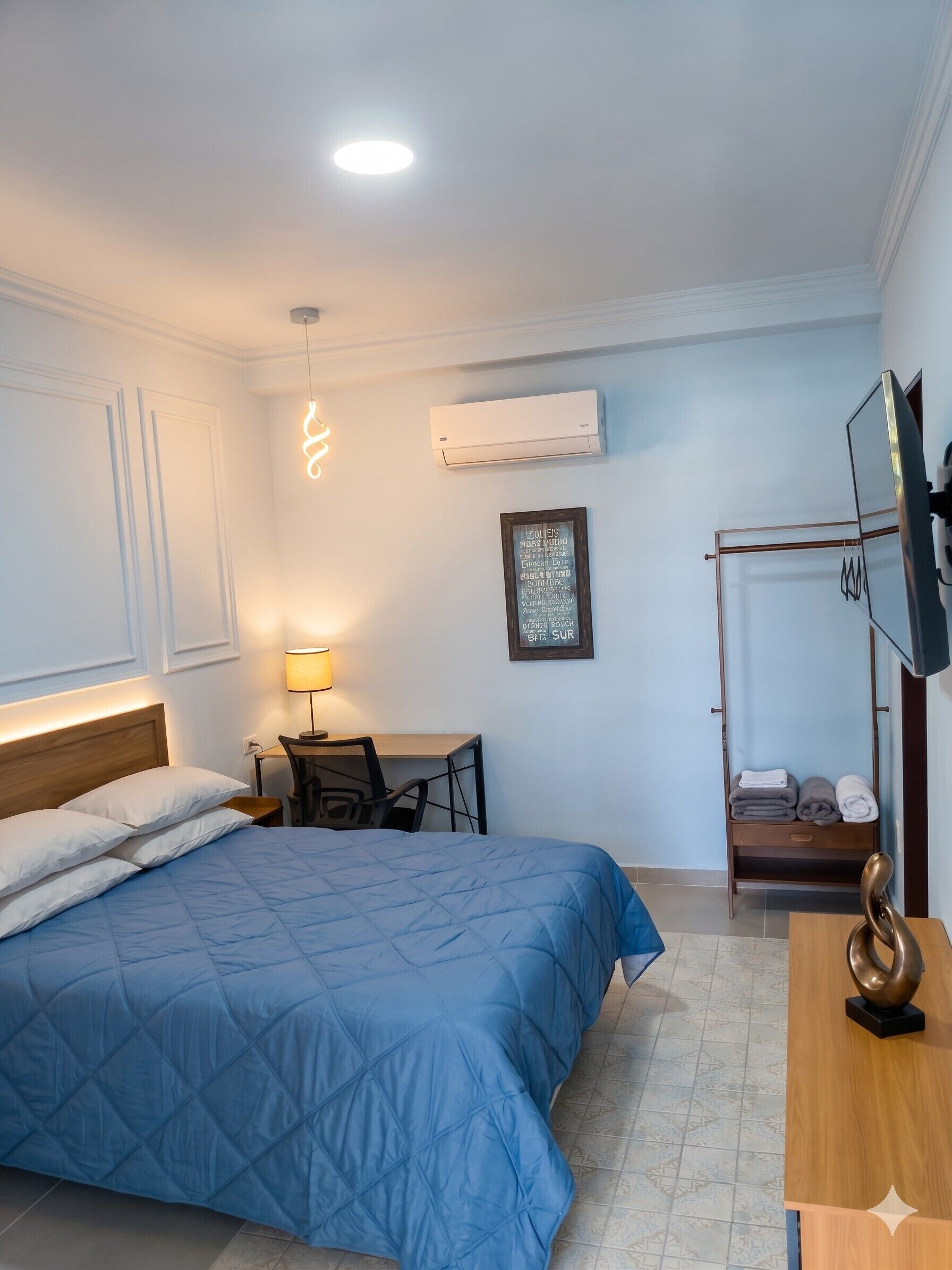 A hotel room with a bed, desk, chair, TV, and a wall-mounted air conditioner.