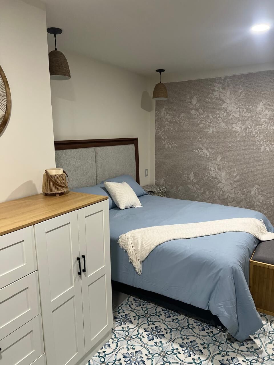 A bedroom with a bed, a wooden nightstand, a wardrobe, and a wall with a floral pattern.