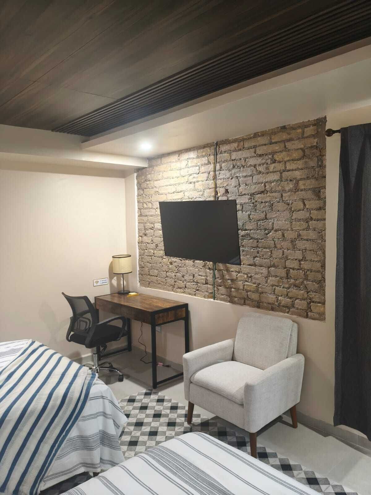 Een hotelkamer met een bed, een bureau, een stoel, een televisie bevestigd aan een bakstenen muur en een geruit vloertje.