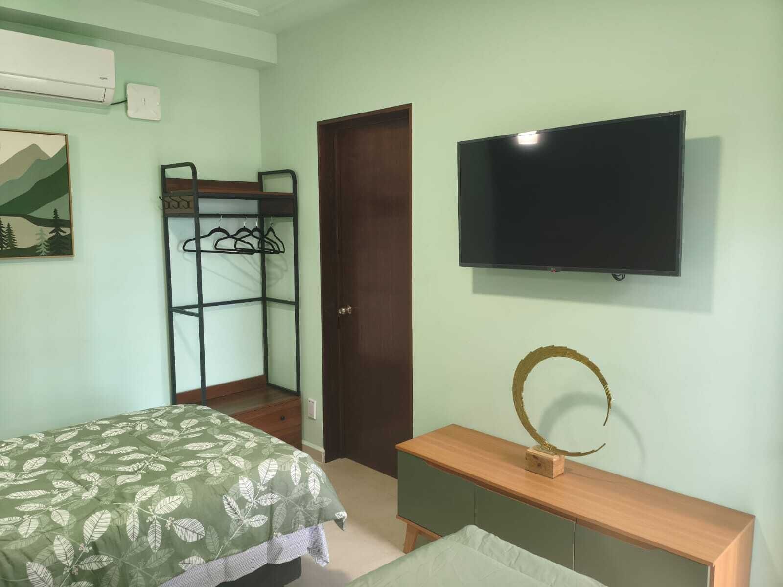 Un dormitorio con una cama, televisión, cómoda de madera, armario con perchas y una puerta.