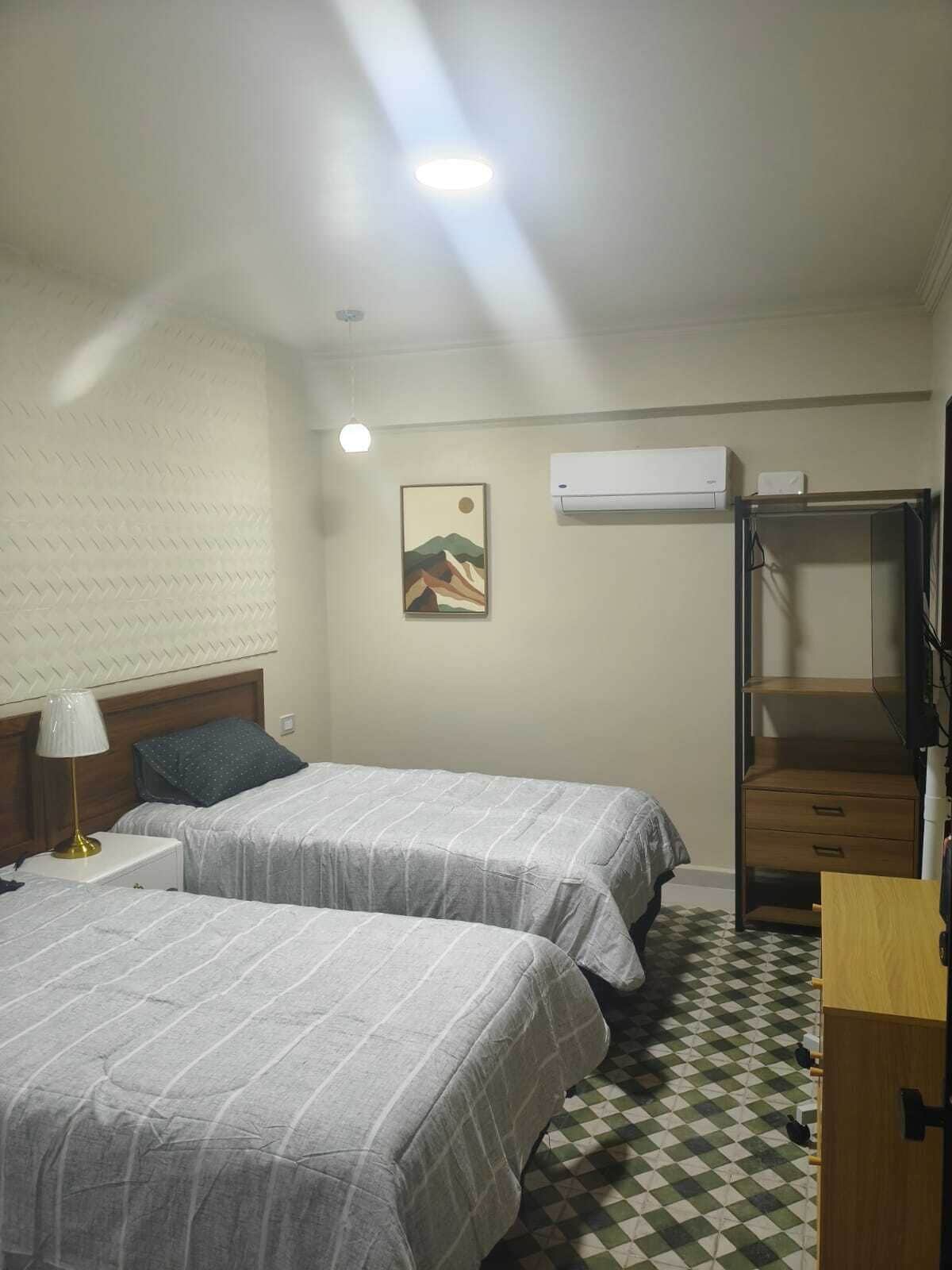 A hotel room with two beds, a nightstand, a lamp, an air conditioner, a painting, and a checkered floor.