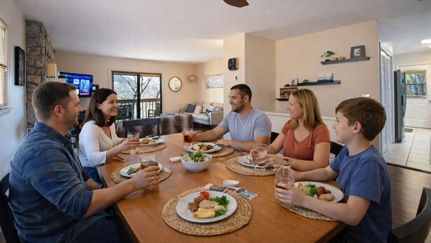 A family dining at a wooden table in a well-lit room with a ceiling fan, a clock, and a television on the wall. - Peaceful 3BR w Hot Tub, in Rumbling Bald, Lake Lure, Resort amenities included (Lake Lure)