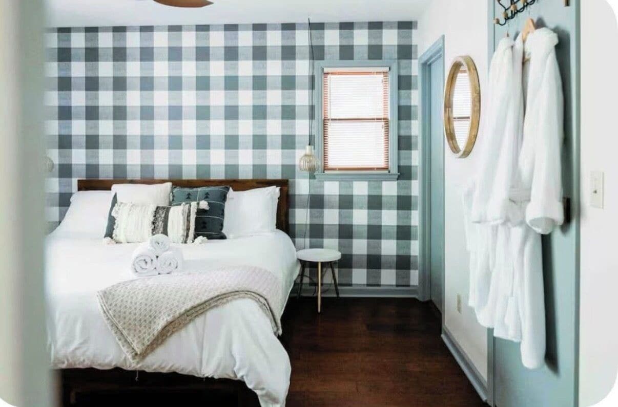 A bedroom with a checkered wall, a bed with pillows, a small stool, and a window with blinds.