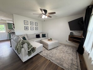 Ein Wohnzimmer mit Ecksofa, Deckenventilator, Flachbildfernseher, einer Küche im Hintergrund und einem großen Teppich.