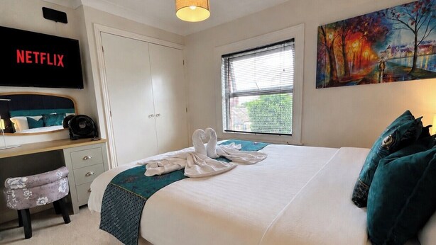 A hotel room with a bed, a desk with a computer, a TV displaying 'NETFLIX', a window with blinds, and a painting on the wall. - Character Bedford Apartment with Two Bedrooms (Bedford)