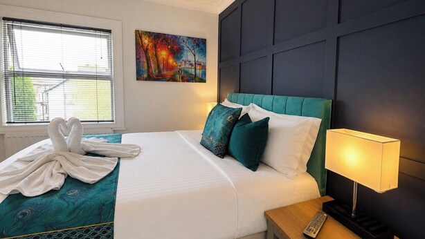 Ein Hotelzimmer mit Bett, einem Bild an der Wand, einem Fenster mit Jalousien und einer Nachttischlampe.