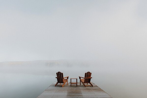 A wooden dock with two Adirondack chairs and a small table, overlooking a foggy landscape.