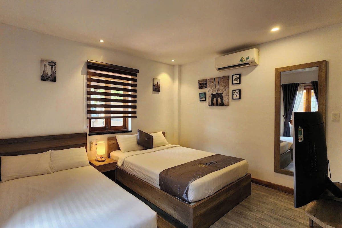 A hotel room with two beds, a window with blinds, a wall-mounted air conditioner, and a mirror.