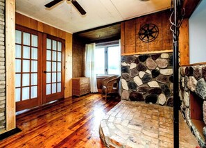 Een kamer met houten vloeren, een stenen open haard, een plafondventilator van hout en een groot raam.