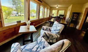Ein Zimmer mit einem großen Fenster, einem Tisch mit einer Vase, zwei gemusterte Stühle, einem Esstisch mit Stühlen und einer Küchenzeile.