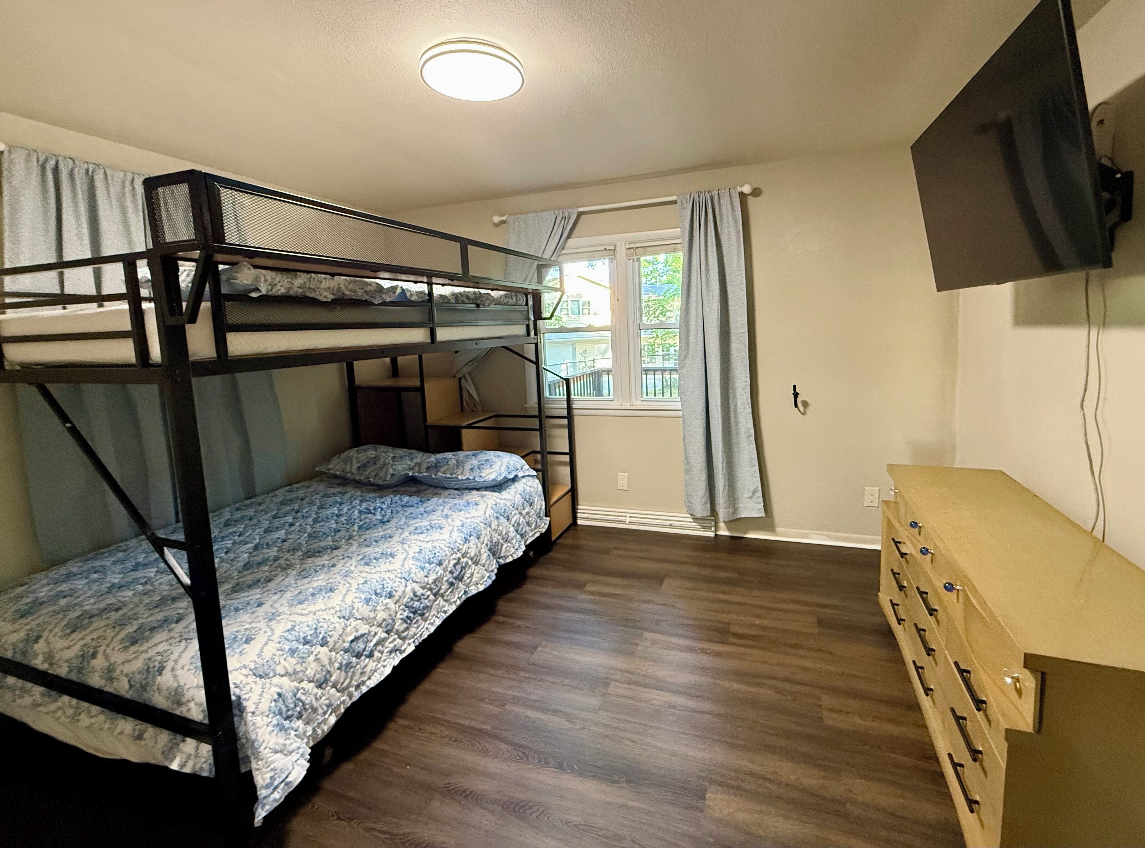 Una camera con letto a castello, pavimento in legno, scrivania con cassetti, finestra con tende e TV montata a parete.