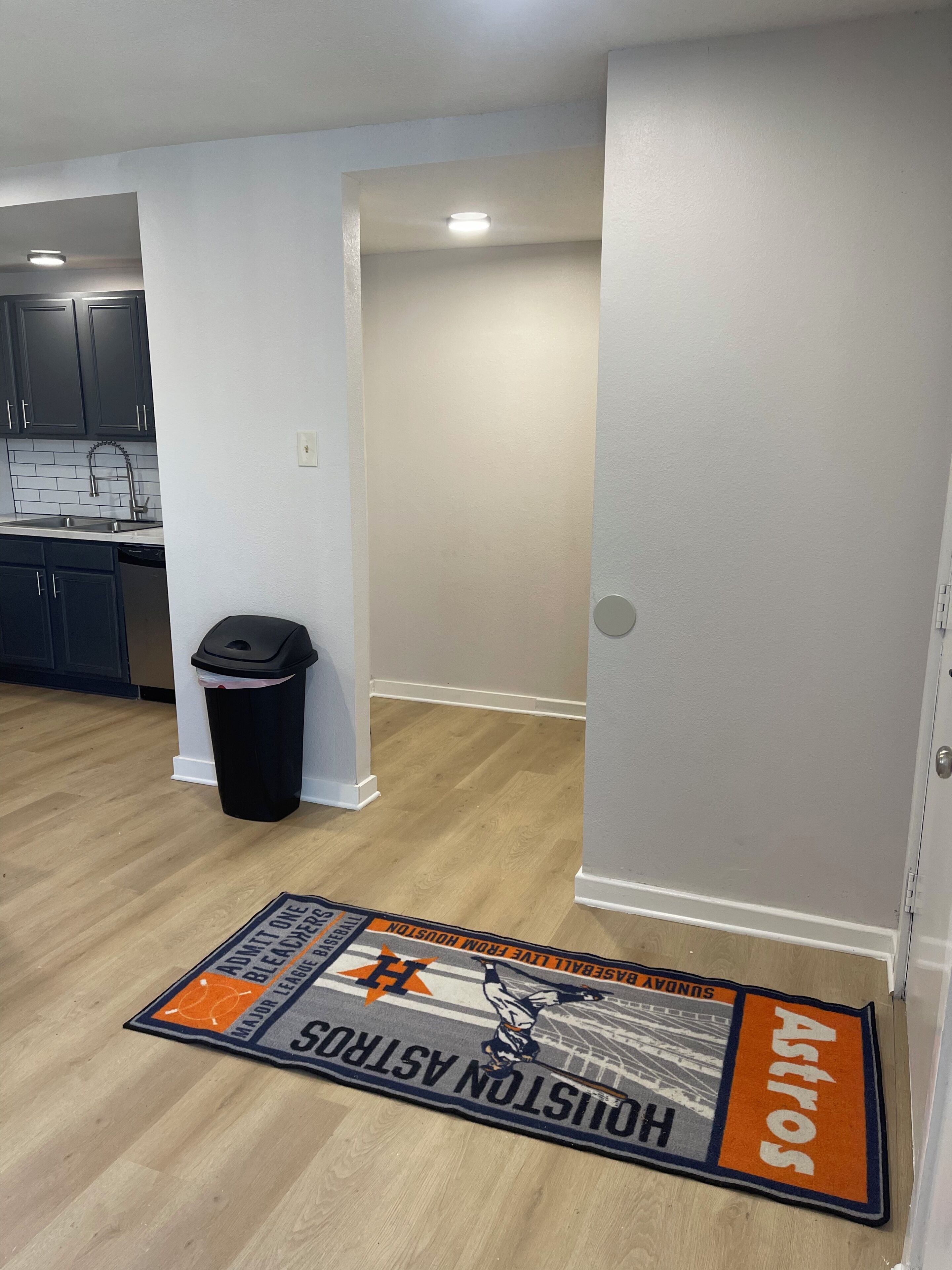 A hallway with a door, a trash can, and a sports-themed mat on the floor.