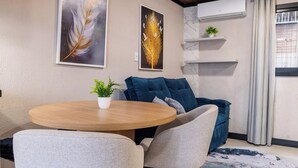 A modern living room with a round wooden table, a blue sofa, and a grey armchair. There are two wall-mounted shelves with decorative items and a potted plant. - Studio for Work and Relaxation | Pool + AC | Pet Friendly | Novo Hamburgo (Novo Hamburgo)