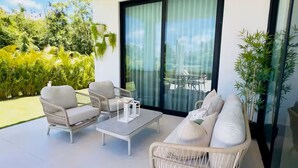 Un espacio exterior moderno con mobiliario de mimbre, un ventanal de vidrio que da a un balcón y vistas al verdor.