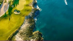 Vista aérea de un campo de golf costero con un camino despejado, agua y el rastro visible de un bote.
