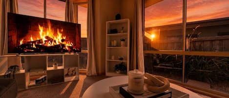 Een woonkamer met een open haard, een televisie en uitzicht op de ondergaande zon door grote ramen.