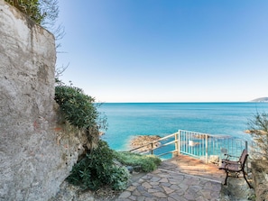 Una escalera de piedra conduce a un balcón con barandilla metálica y vistas a un mar azul y despejado.