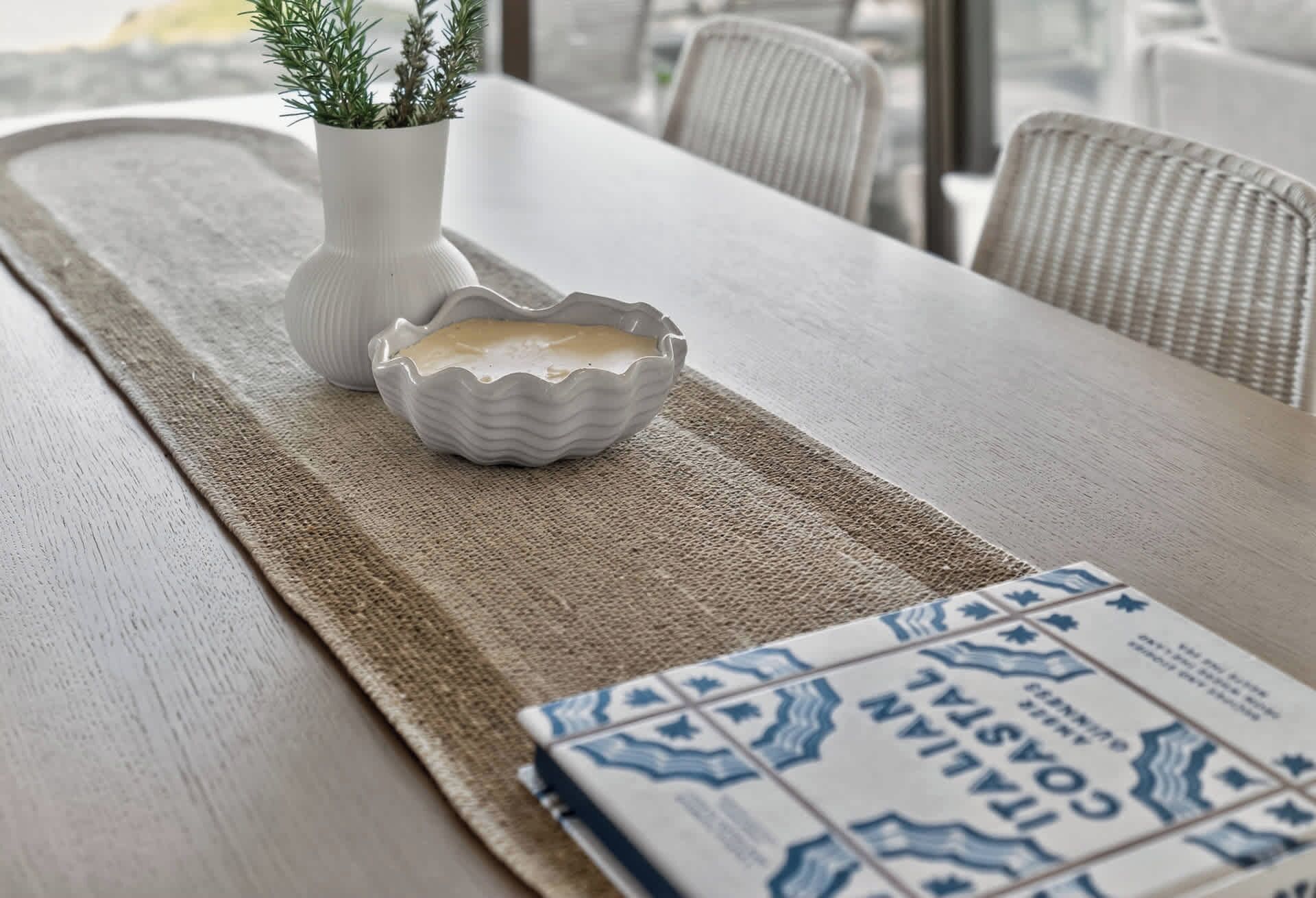 A dining table with a placemat, a white ceramic bowl, a white vase with greenery, and a magazine.