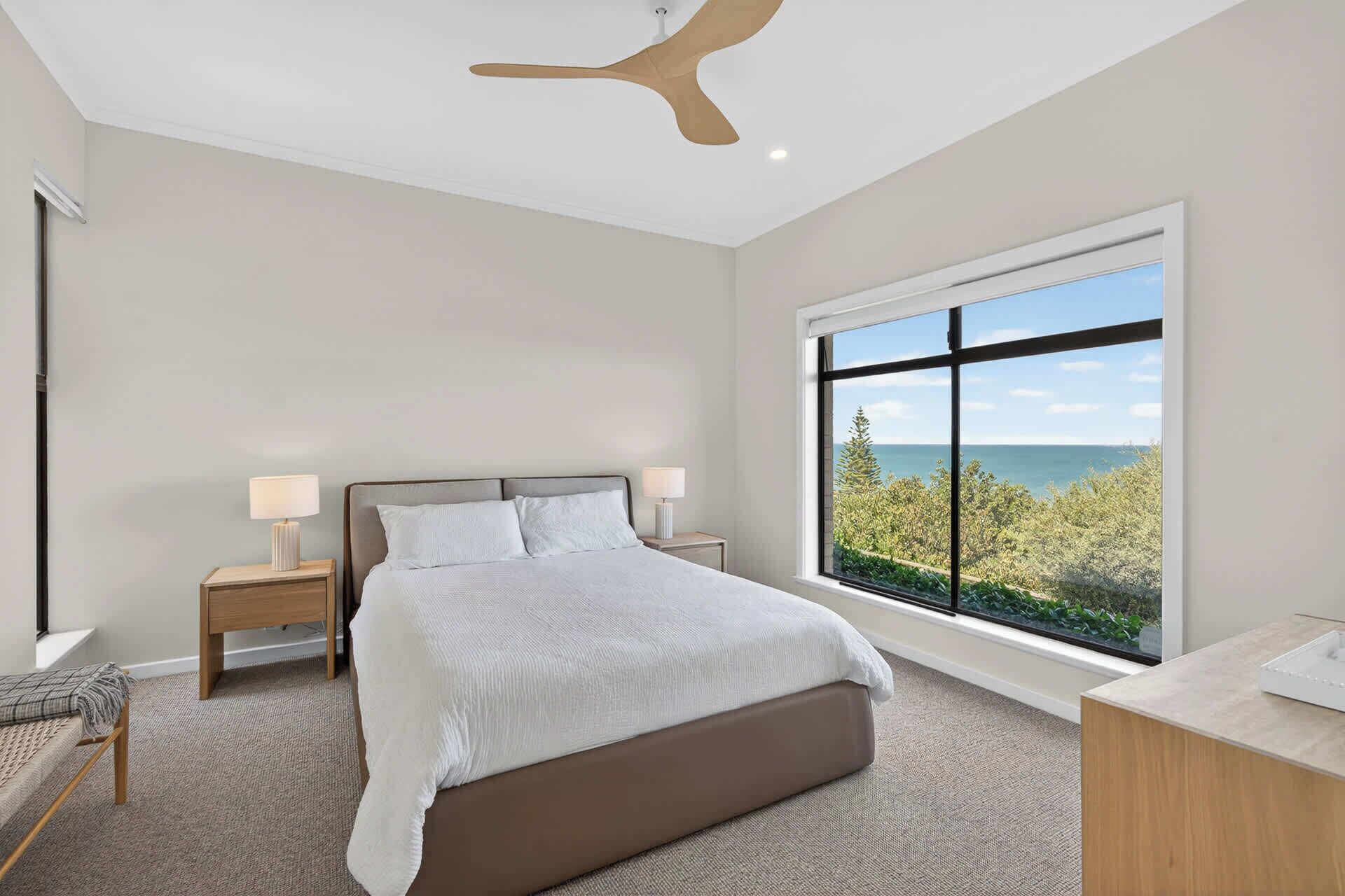 A bedroom with a bed, two bedside tables with lamps, a window with a view of the sea, and a ceiling fan.