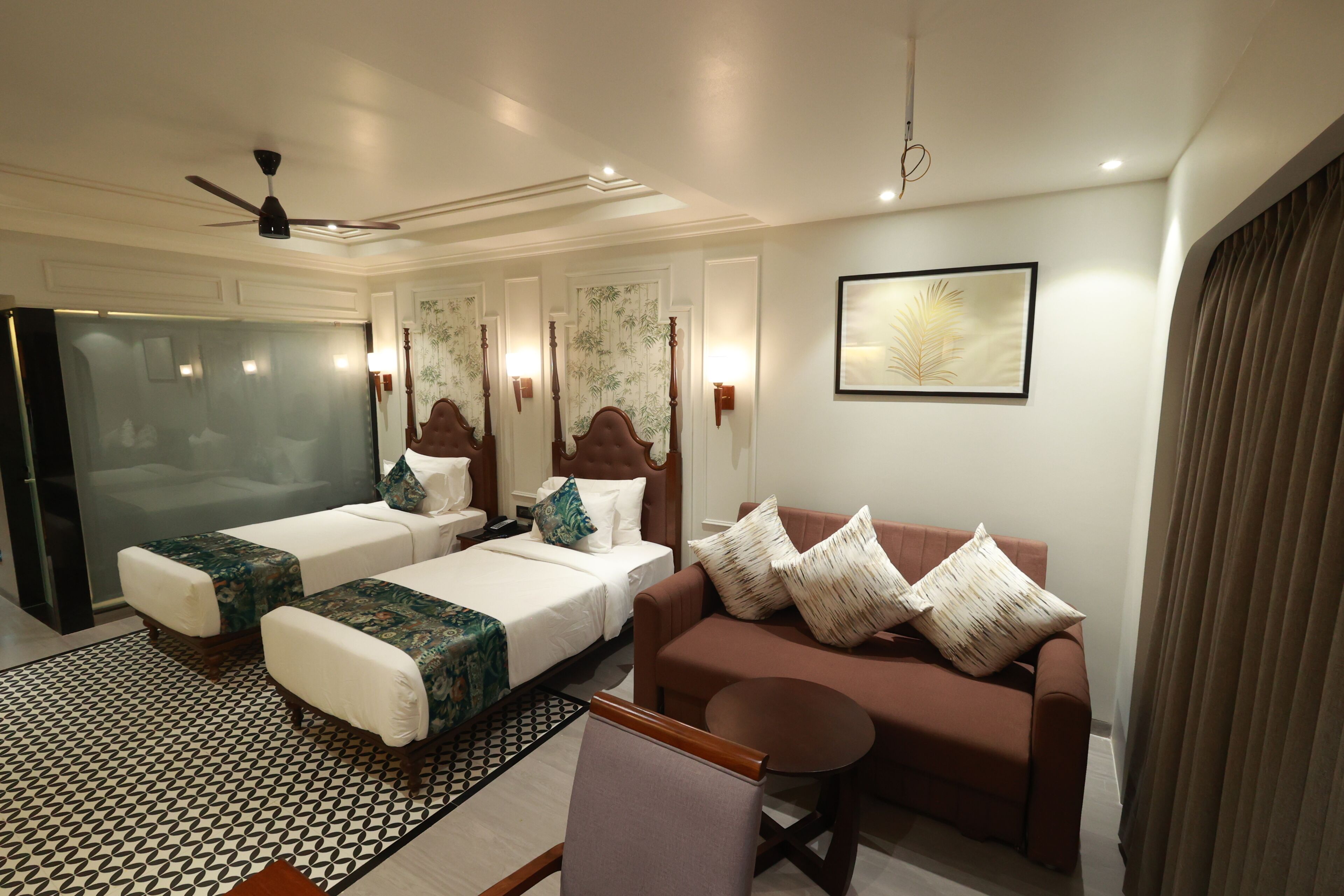 A hotel room with two single beds, a sofa, a small table, a ceiling fan, and a large window.