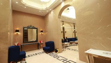 Um hall de hotel com piso de mármore, um espelho grande, duas poltronas azuis, uma mesinha e um console decorativo.