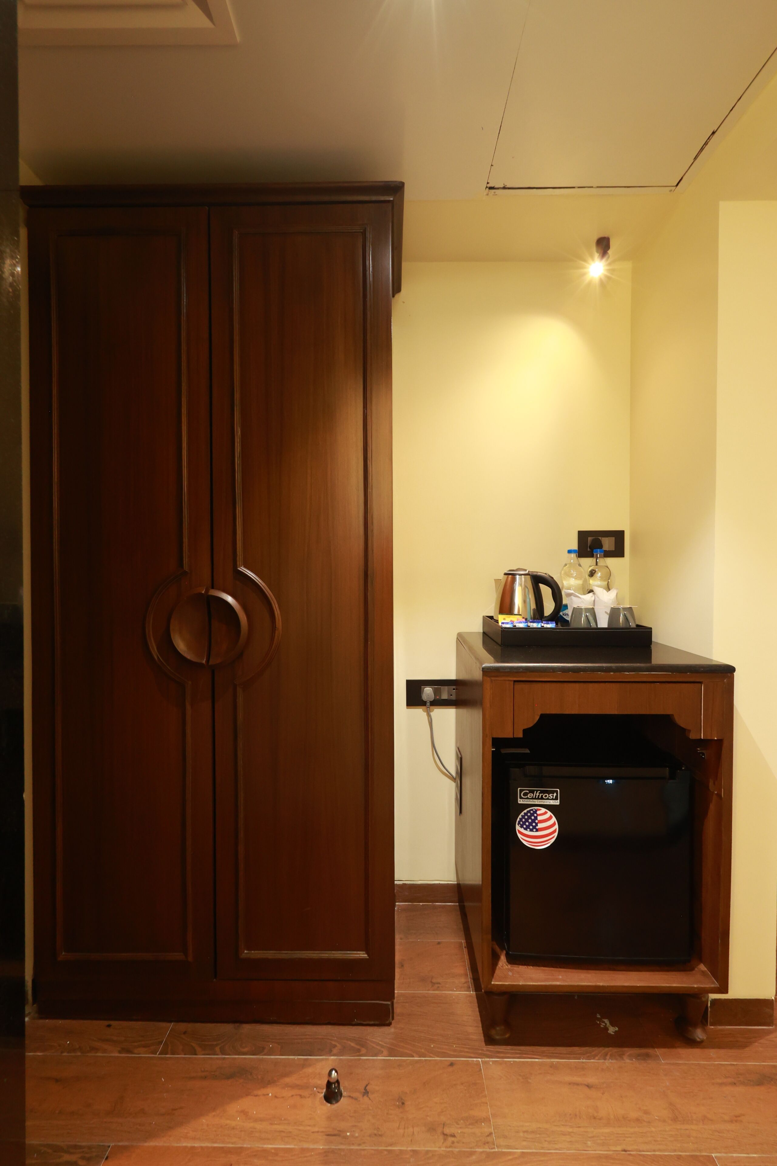 A room with a wooden wardrobe, a black refrigerator with a sticker, and a small table with a kettle and a bottle.