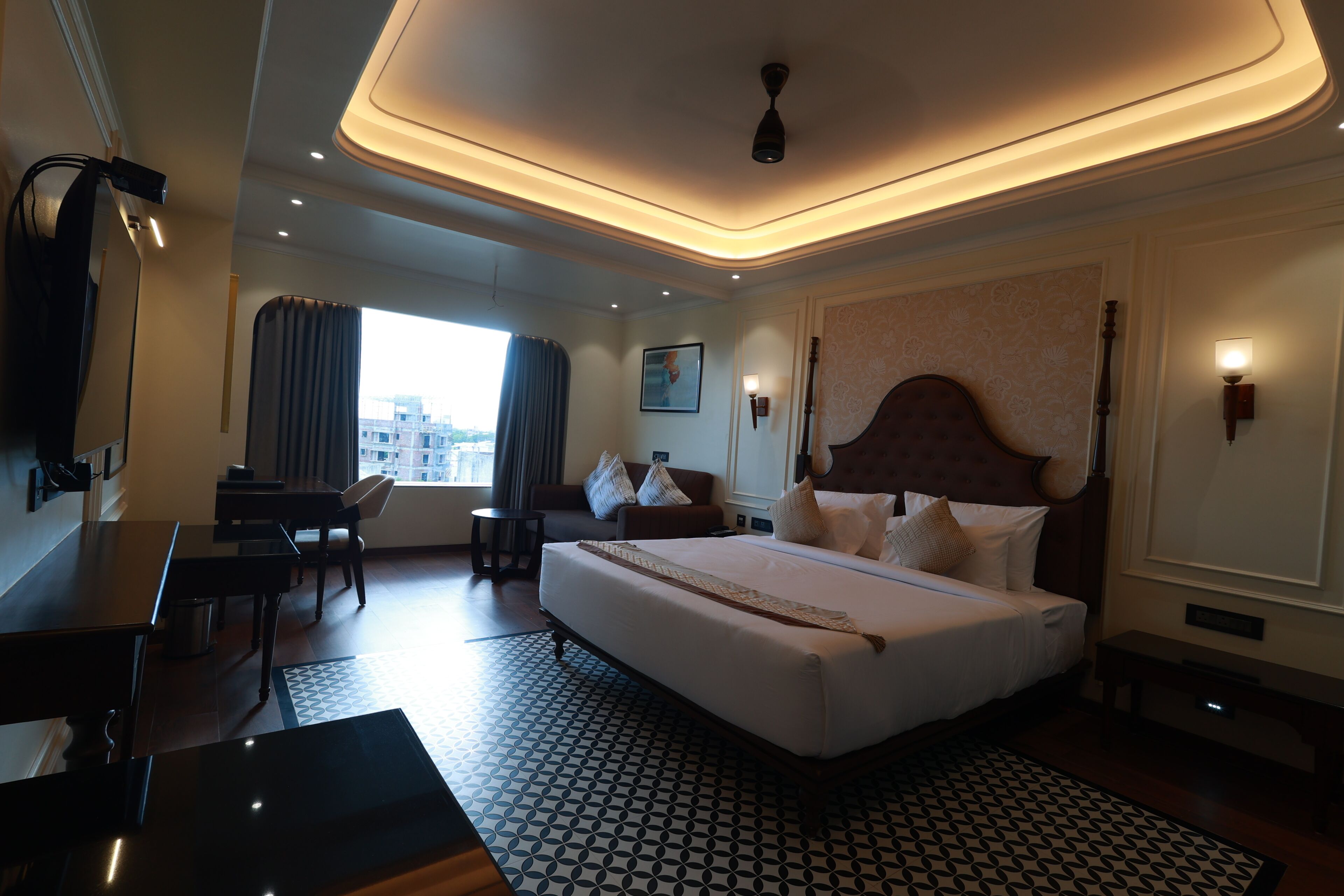 Quarto de hotel com uma cama grande, uma escrivaninha, uma cadeira, uma mesa pequena, uma luminária no teto e uma janela com cortinas.