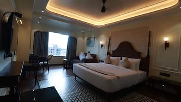 Quarto de hotel com uma cama grande, uma escrivaninha, uma cadeira, uma mesa pequena, uma luminária no teto e uma janela com cortinas.
