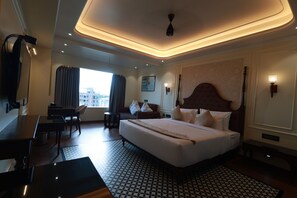 A hotel room with a large bed, a desk, a chair, a small table, a ceiling light, and a window with curtains. - Aurtus (Amravati)