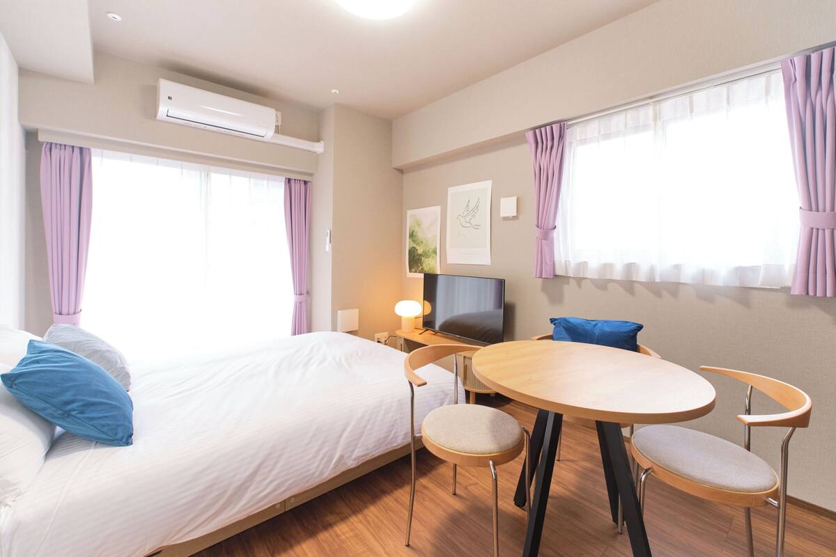 A hotel room with a bed, a desk with a TV, a round table with chairs, and large windows with curtains.