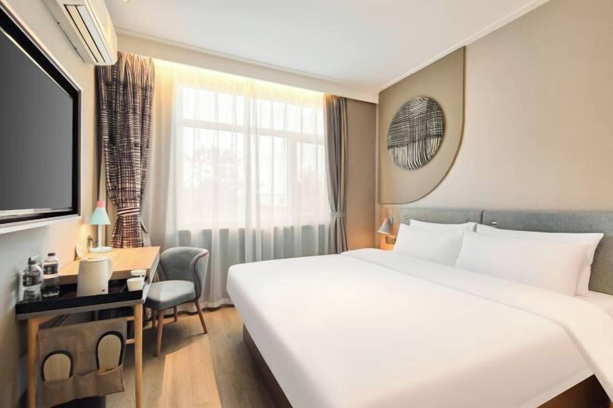 Hotellihuone, jossa on sänky, työpöytä, tuoli ja suuri ikkuna, jossa on verhot.