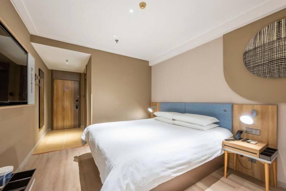 Hotellihuone, jossa on sänky, yöpöytä, lamppu ja seinään kiinnitetty valaisin.