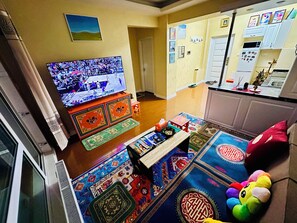 A living room with a television, a small table, colorful rugs, and a kitchen area with cabinets and a refrigerator.