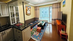 Un espacio habitable compacto con kitchenette, zona de comedor con alfombra colorida, sofá, televisor y ventana grande con cortinas.