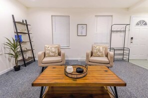 Een kamer met twee beige fauteuils, een houten salontafel, een kleine plant in pot, een metalen plank en een deur met raam.