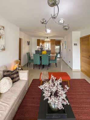 A living room with a sofa, a coffee table with flowers, a dining area with patterned chairs, and a kitchen in the background.