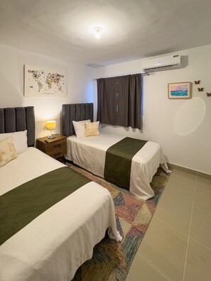 A hotel room with two beds, a nightstand, a lamp, a world map artwork, a window with curtains, an air conditioning unit, and a small framed picture.