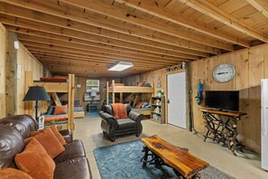 Un accogliente interno di una baita con soffitto in legno, un divano, una poltrona, un tavolo, una televisione e una libreria.