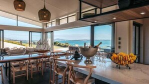 A modern dining area with a view of the sea, wooden furniture, and a bowl of fruit on the counter. - Tradewinds Retreat by Waiheke Pure - Views views views, pool, spa, luxurious spacious living (Waiheke Island)