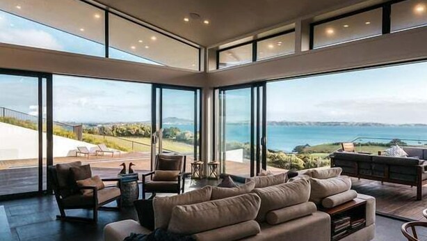 A spacious living area with large windows, a comfortable sofa, and a view of the sea. - Tradewinds Retreat by Waiheke Pure - Views views views, pool, spa, luxurious spacious living (Waiheke Island)